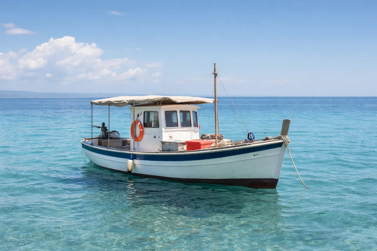 Small white fishing boat anchored on calm turquoise sea, showing a simple cabin, lifebuoy, clear water, and a peaceful coastal background.