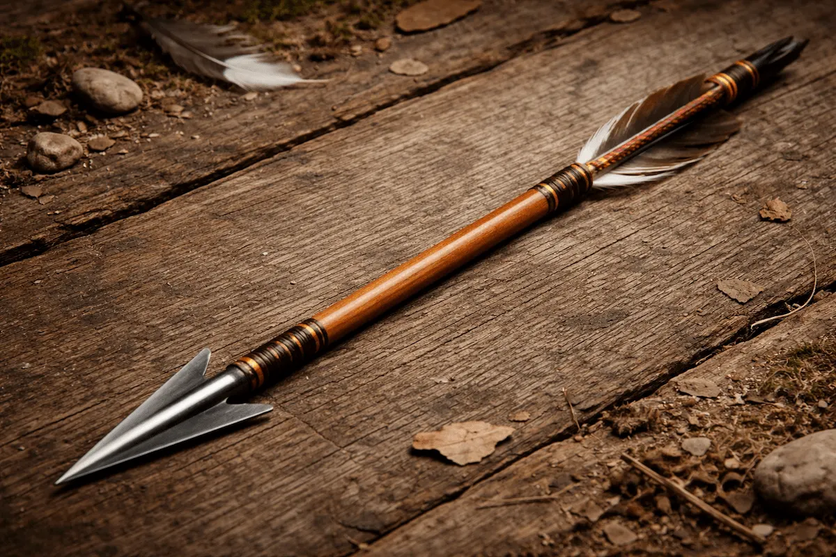Wooden arrow with a metal tip and feather fletching lying on a weathered wooden background, showing traditional archery equipment.